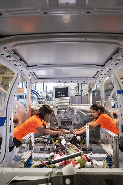 Two female automotive engineering students in orange shirts connect electrical wires controlling a screen in a vehicle frame.