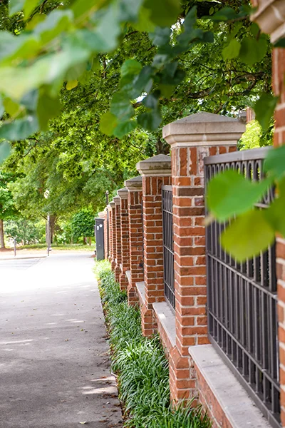 A metal fence with brick columns stretches along a sidewalk lined with trees.