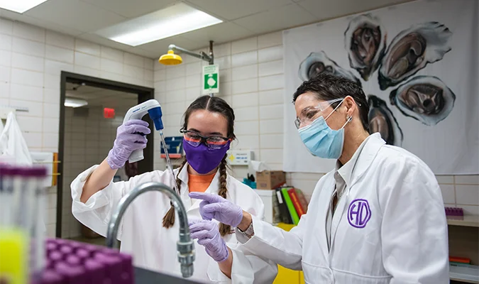A student and professor in lab gear measure samples with a pipet.