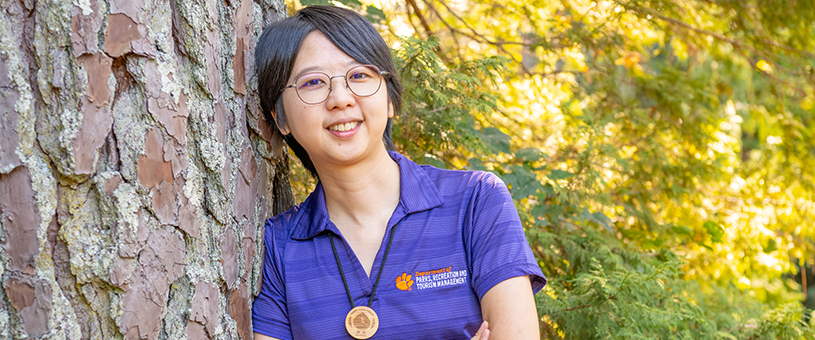 Headshot of Hsuan Hsieh leaning against a tree with a forest in the background.