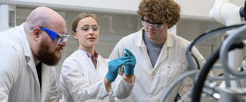Three researchers observe a beaker together in a laboratory setting.