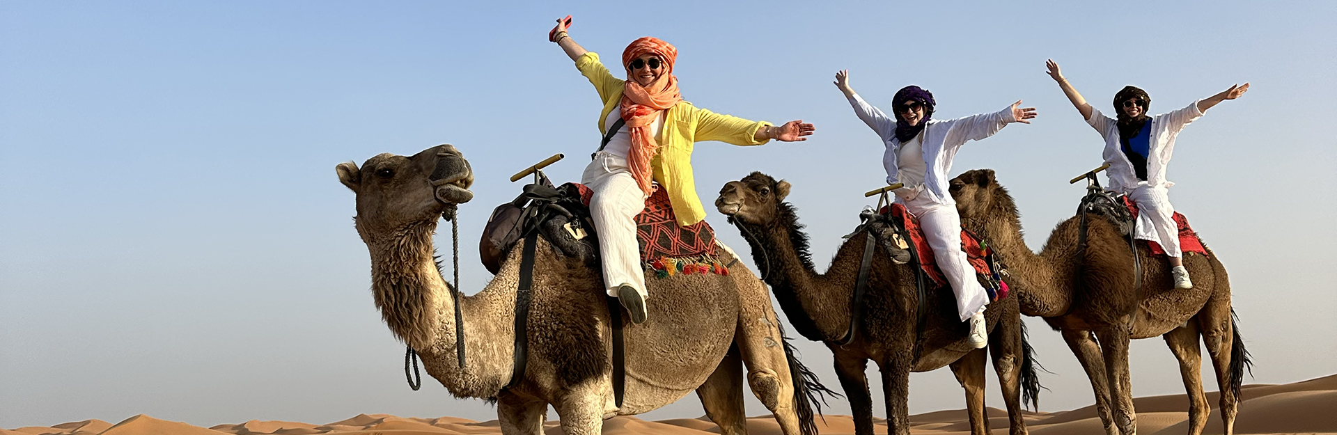Three people in the desert on top of camels posing with their arms stretched out toward the sky.