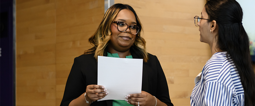 Two women in professional attire chat while holding paperwork in their arms