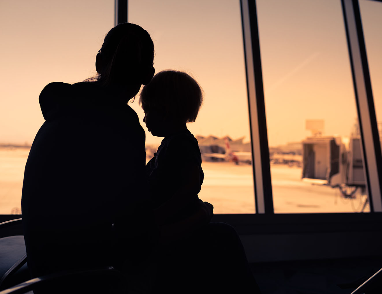 A parent and child await travel in an airport in front of a backlit sunset window.