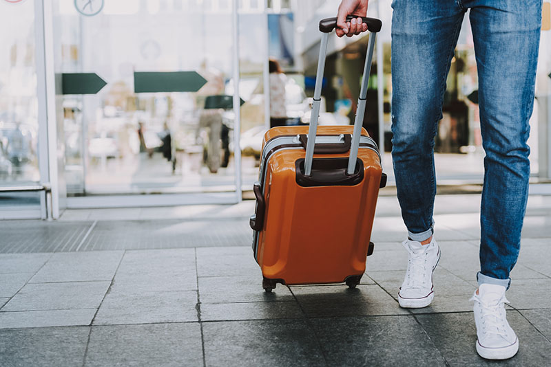 A traveler walking with an orange suitcase at an airport entrance.