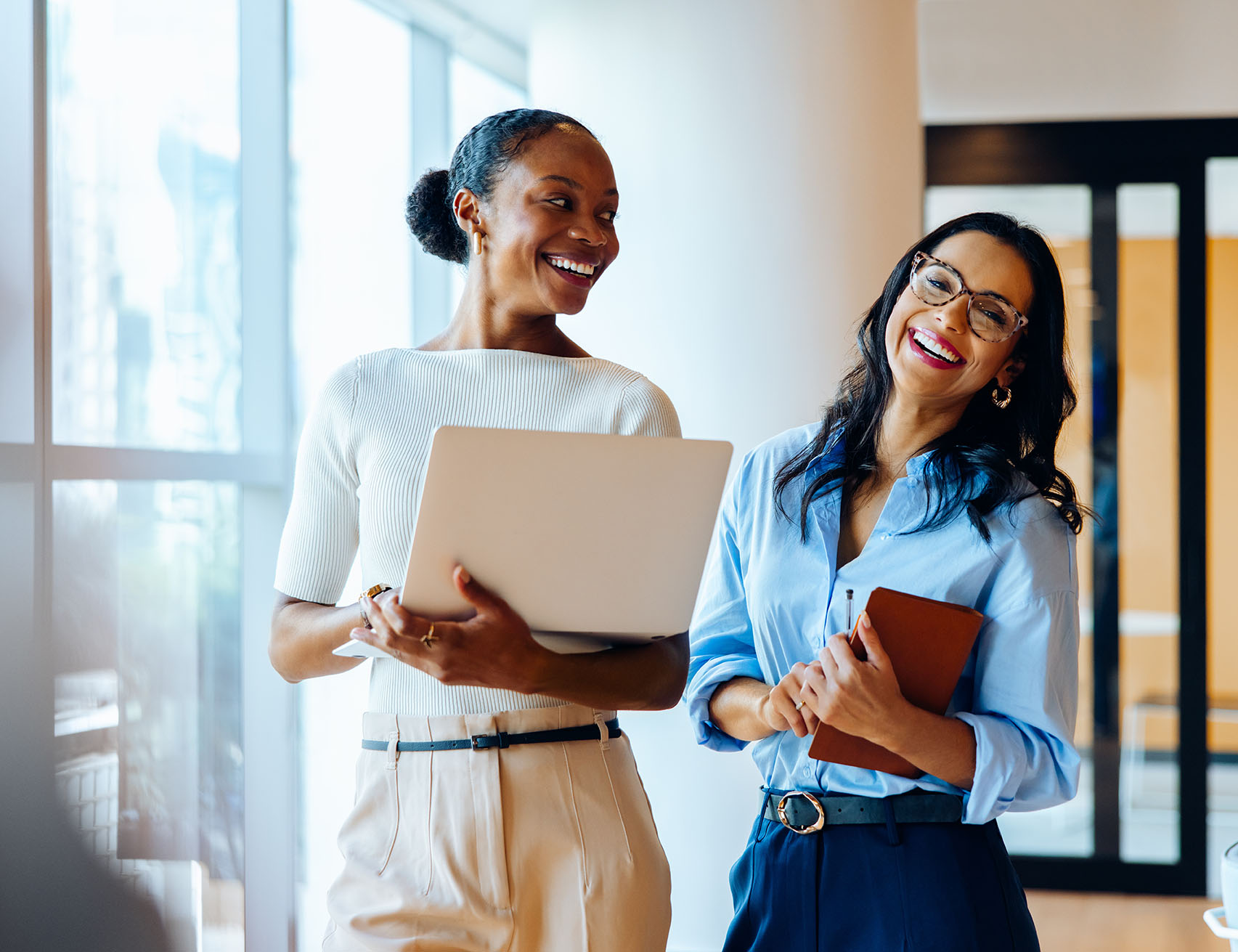 Two smiling women in business attire, one holding a laptop and the other a notebook, engaged in a friendly conversation in a modern office setting.