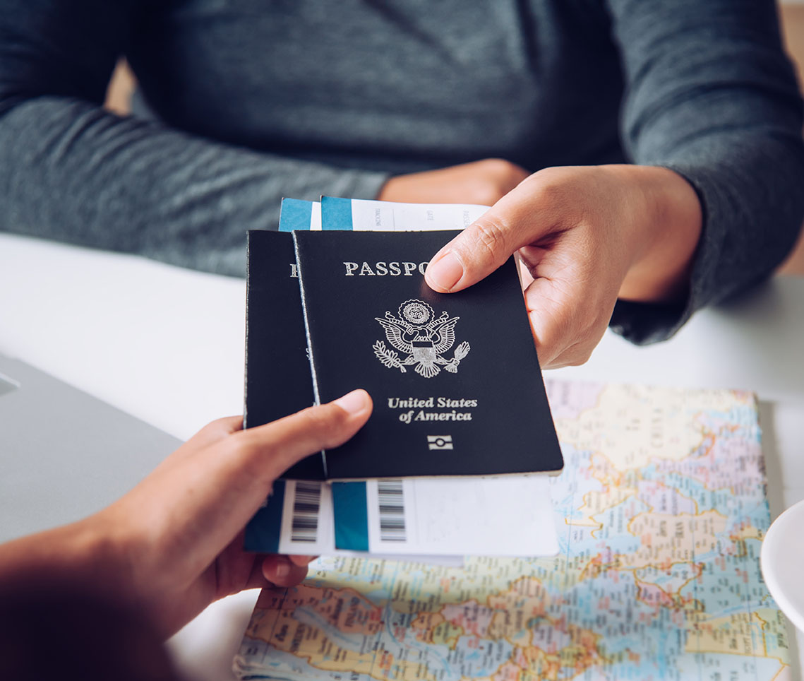 A person's hand receiving a U.S. passport along with travel documents, with a map and a laptop visible on the table.