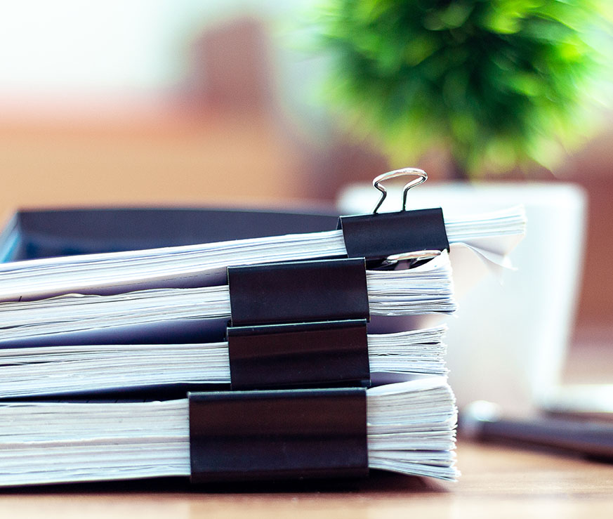 A stack of papers held together with binder clips, placed on a wooden surface with a small potted plant and mug in the background.