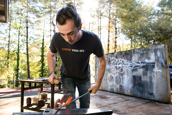 A student leaning over an anvil holding a glowing hot metal rod that they are shaping.