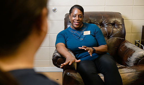 A woman wearing a name badge sits in a leather chair talks to a person just out of view. 