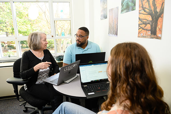 A woman and man look at each other from while talking to a person sitting across the table from them. Each person has a laptop on the table. 