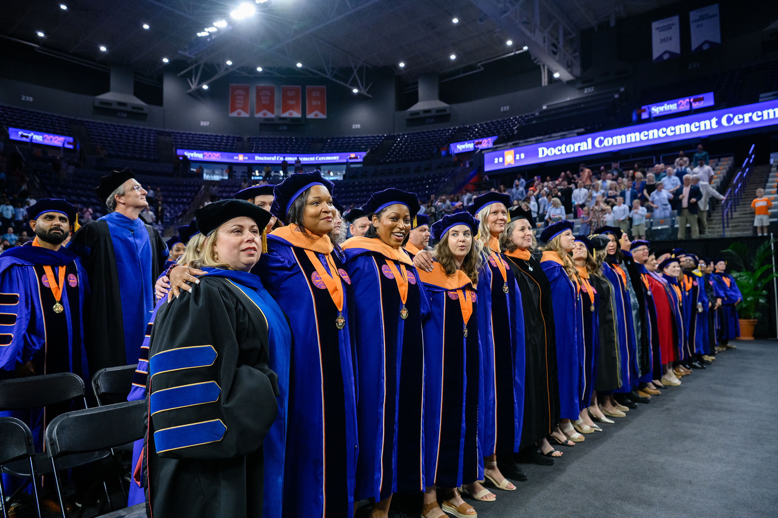 Line of doctoral graduates standing with arms on each other's shoulders.