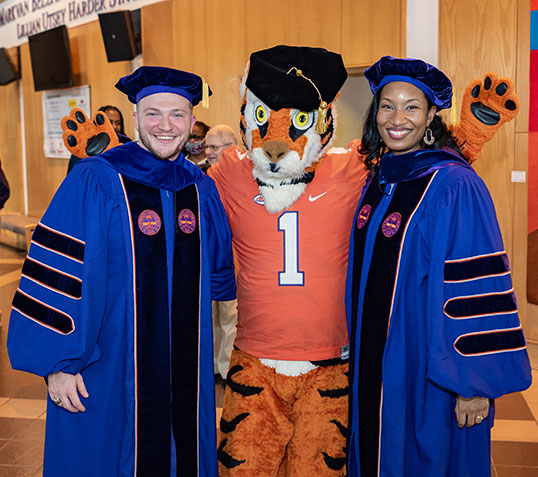 The Clemson Tiger wears a Graduation Tam and poses with two doctoral students wearing their regalia.
