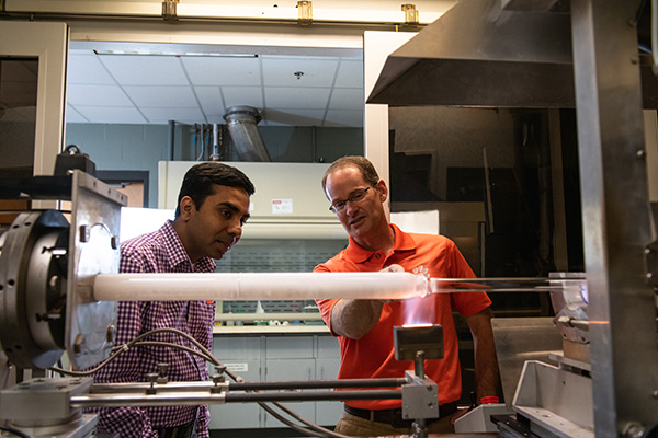 Two men look at a machine heating up a glass-like tube for fiber optics. 