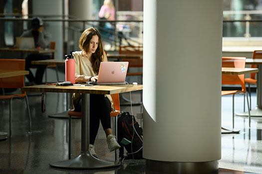 A female student works on her laptop at a small table in the School of Business building.