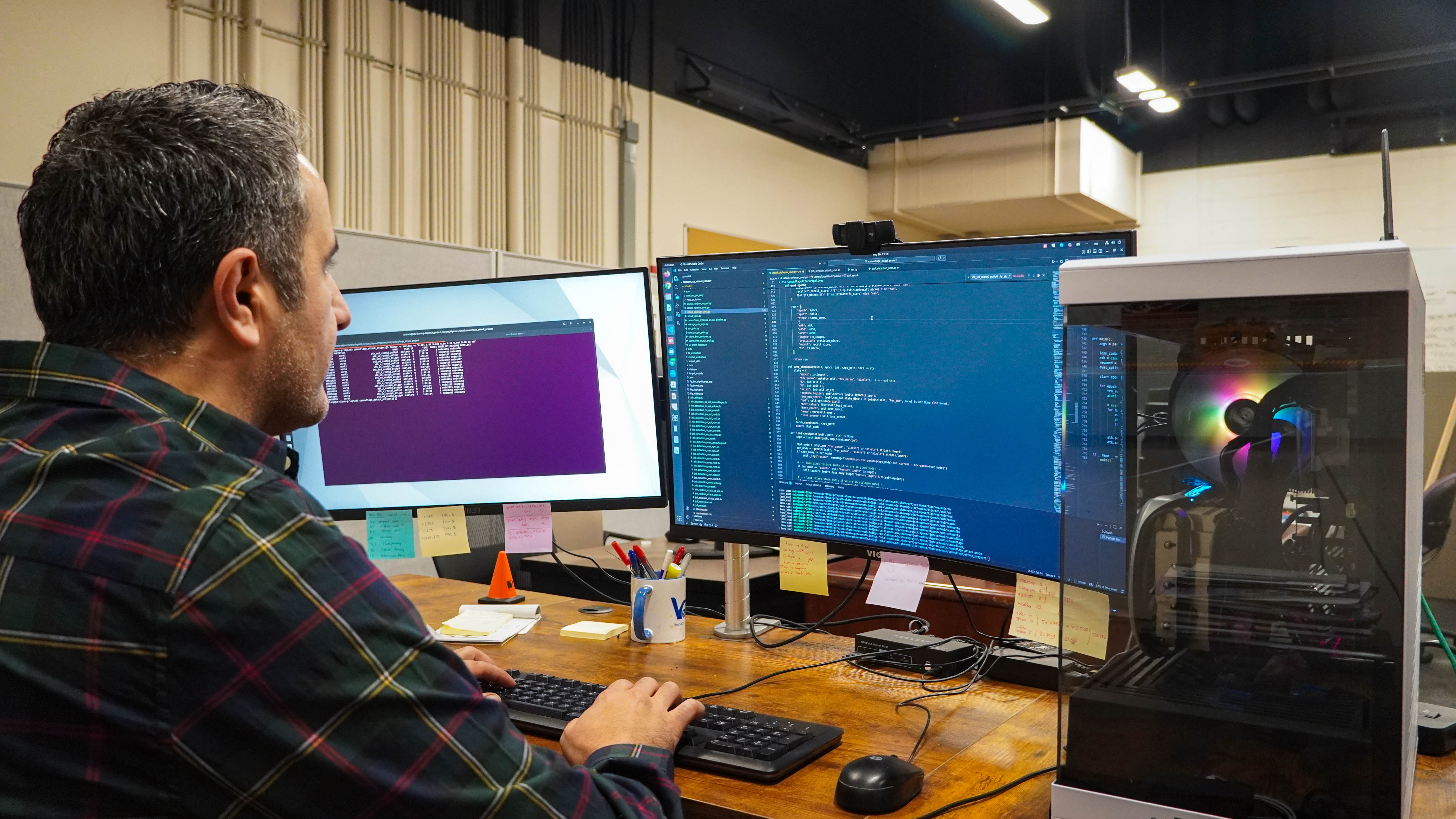 Dr. Amir Salarpour seated at a desk in an office, using a keyboard and mouse beside dual monitors showing code and an open desktop computer with colorful internal lights.