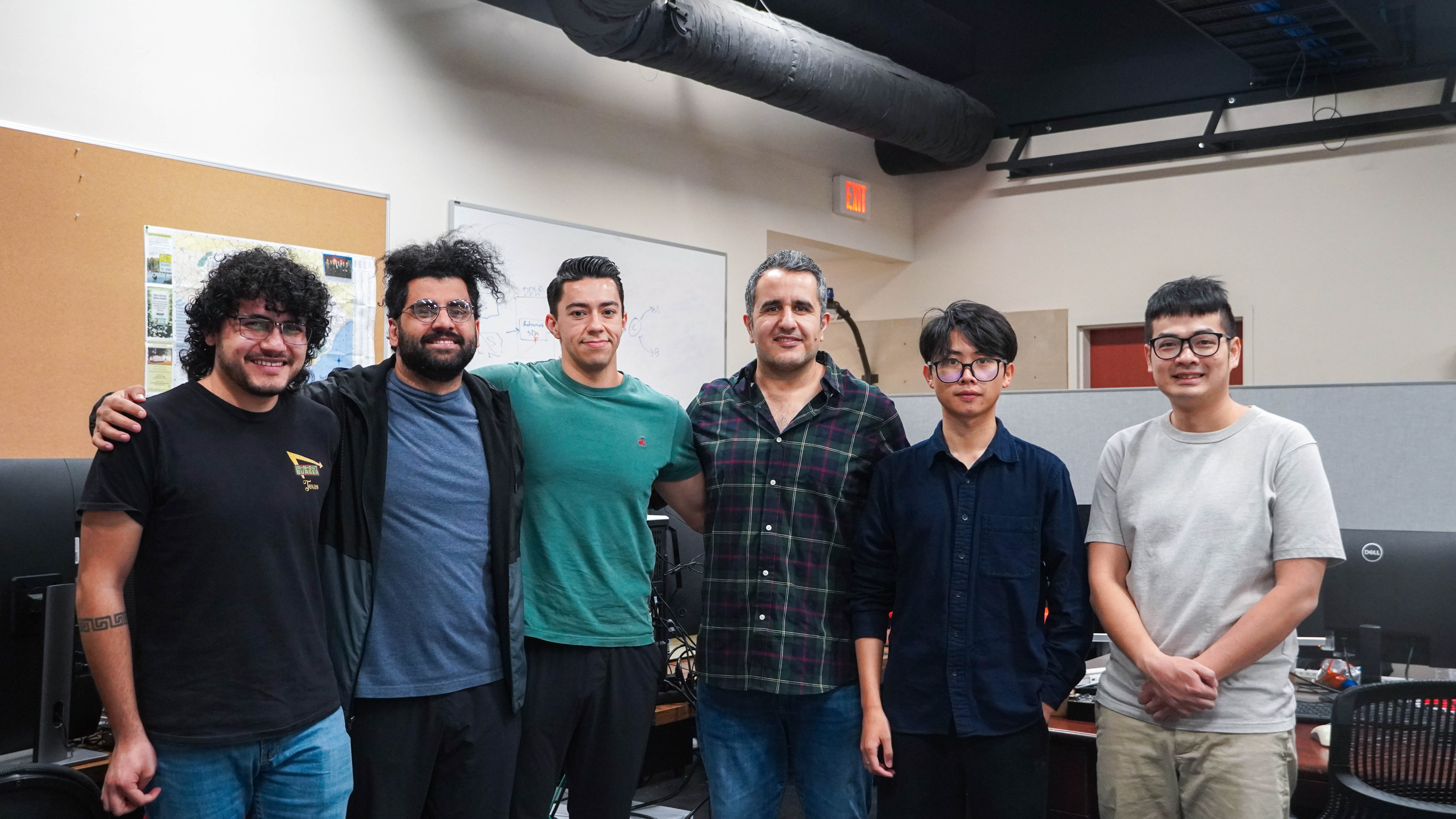 Six colleagues standing side-by-side in an open office, with Dr. Amir Salarpour featured third from the right wearing a dark plaid shirt. Computer monitors and a whiteboard are visible in the background.