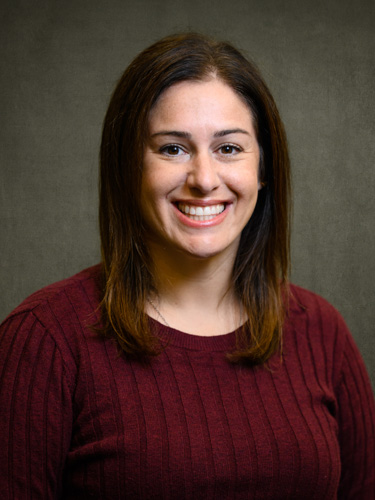 Smiling woman with shoulder-length brown hair wearing a maroon sweater against a gray studio backdrop.