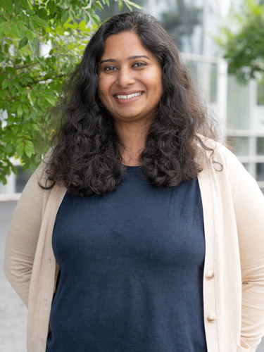 Smiling woman with long, curly dark hair standing outdoors near leafy greenery, wearing a navy top and light cardigan.