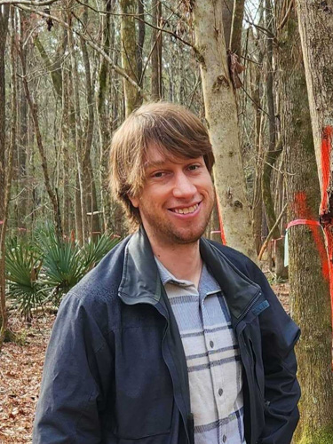 Smiling man in a jacket standing in a wooded area with leafless trees, including a tree marked with red paint.