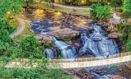 Aerial view of the illuminated Liberty Bridge spanning over the Reedy River Falls at dusk in downtown Greenville, South Carolina.