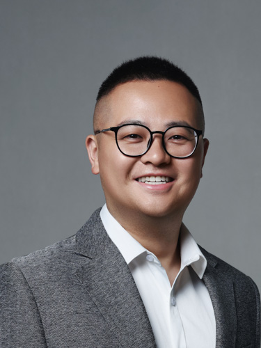 Smiling person wearing round glasses, a gray blazer, and a white shirt against a plain gray background.