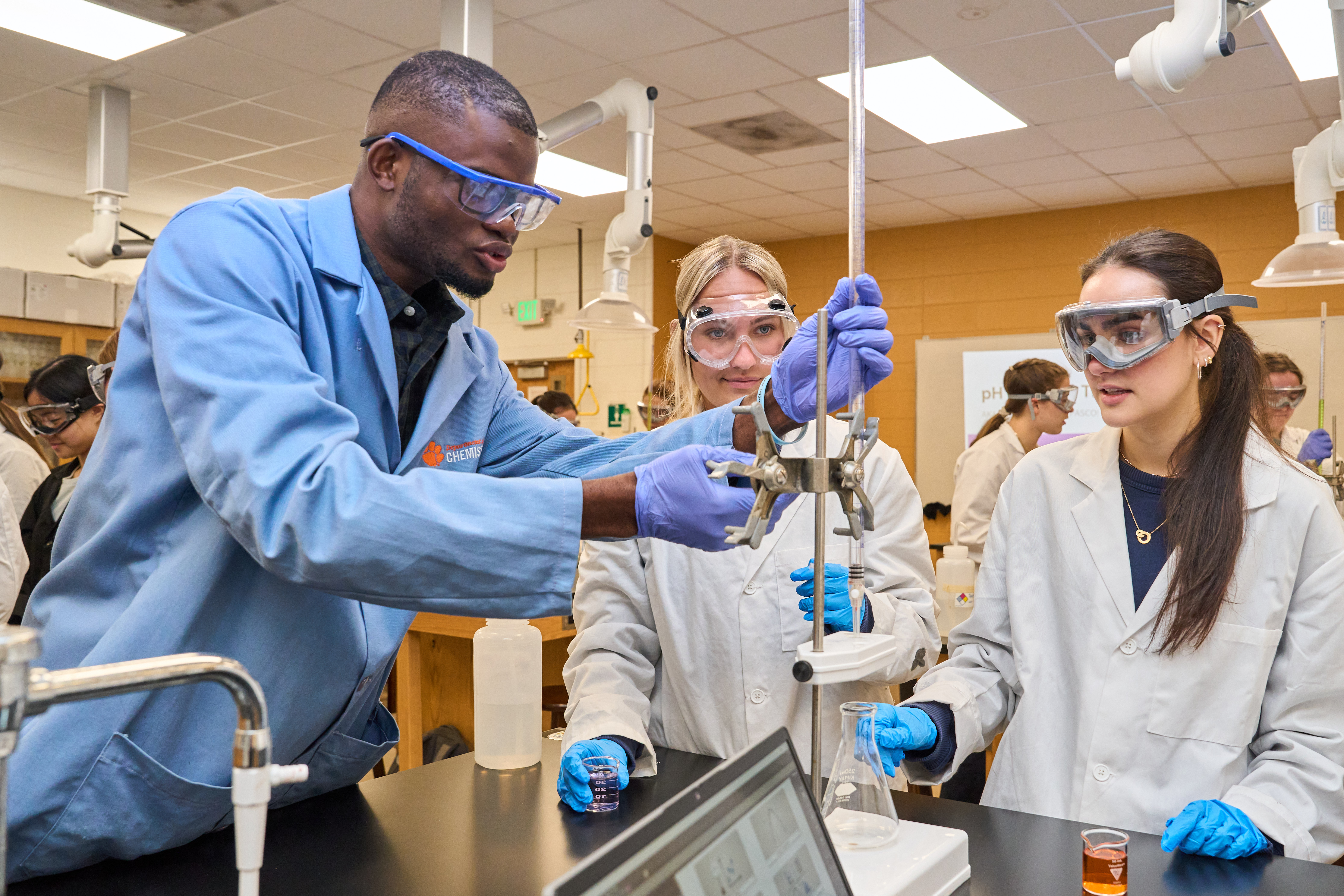 Instructor in a chemistry lab demonstrates a measurement setup while students in lab coats, gloves, and safety goggles watch at a lab bench.