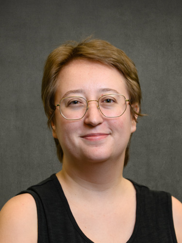 Studio-style headshot of a person with short light-brown hair and round glasses, smiling against a gray background.
