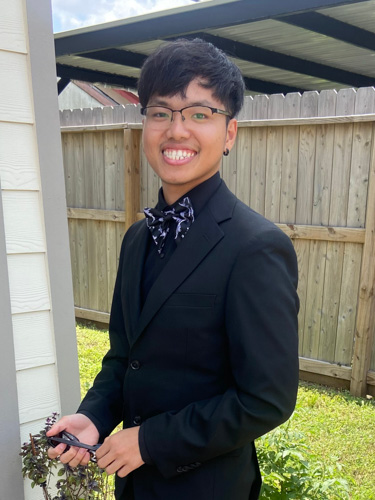 Smiling person in a black suit with a patterned bow tie, standing outdoors by a wooden fence and holding a phone.