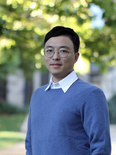 Portrait of a person wearing round glasses and a blue sweater over a white collared shirt, standing outdoors with blurred green trees in the background.