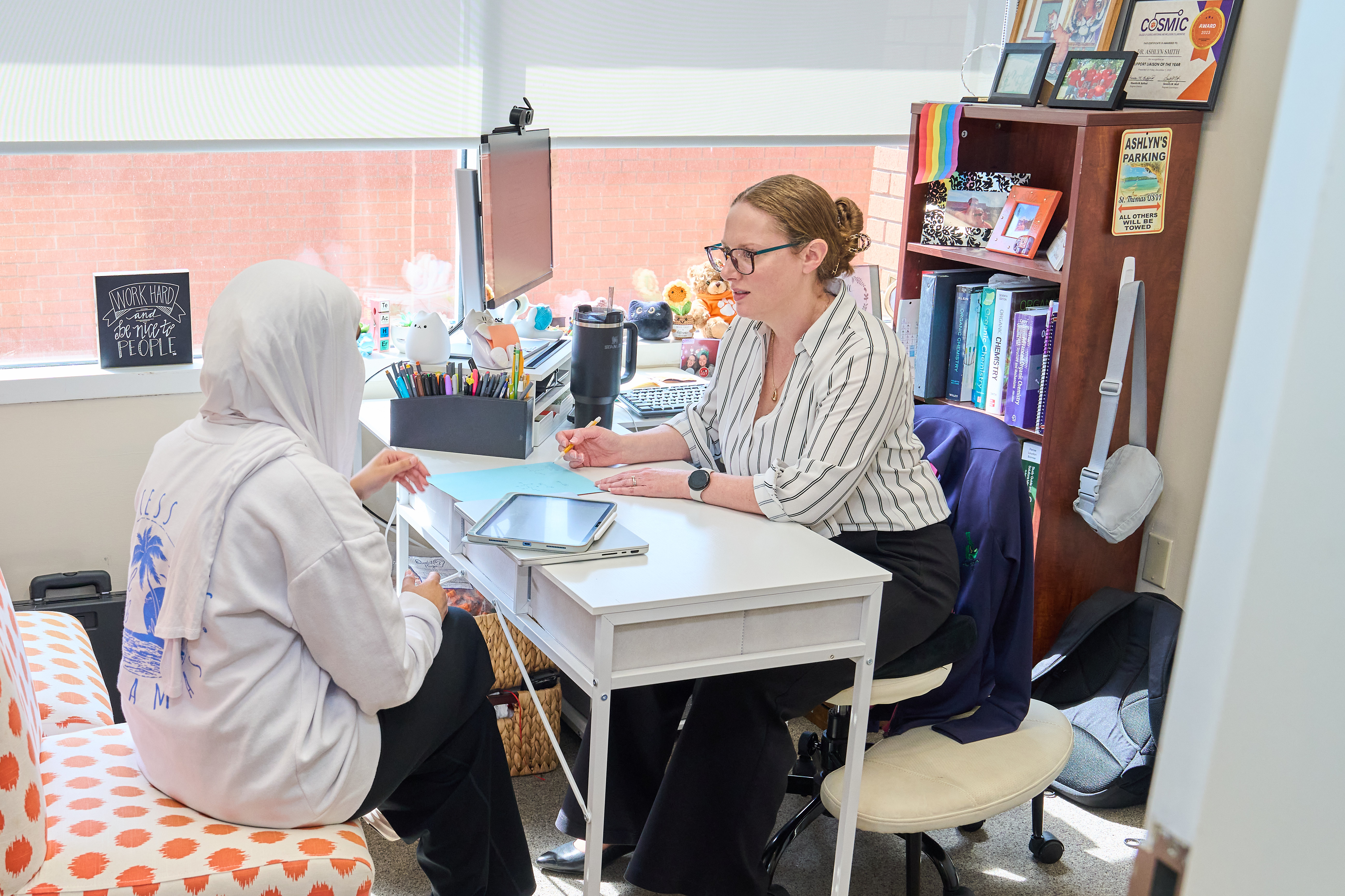 Teacher and student talk across a desk in a school office, with books and photos on a shelf nearby.