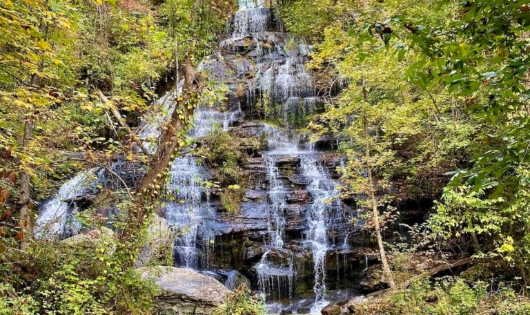 A wide, multi-tiered waterfall cascading down dark, rocky ledges surrounded by dense green and yellow autumn trees.