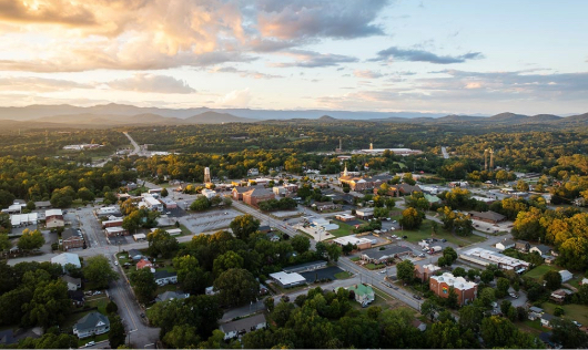 Aerial sunset view of downtown Pickens, South Carolina, showing city streets nestled in dense green trees with the Blue Ridge Mountains in the distance.