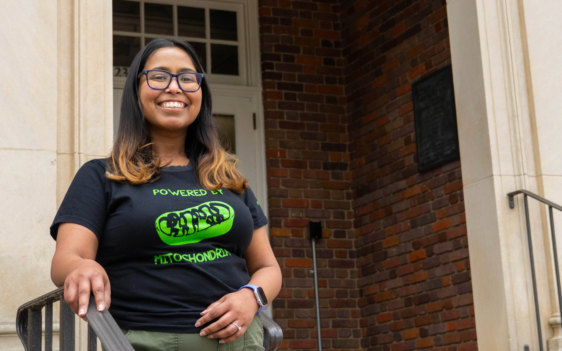 Dr. Pro Manna wearing glasses and a black “Powered by mitochondria” T-shirt stands on steps outside a brick building.