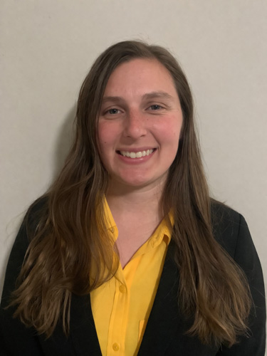 Head-and-shoulders portrait of a smiling person wearing a yellow shirt and black blazer against a plain light background.