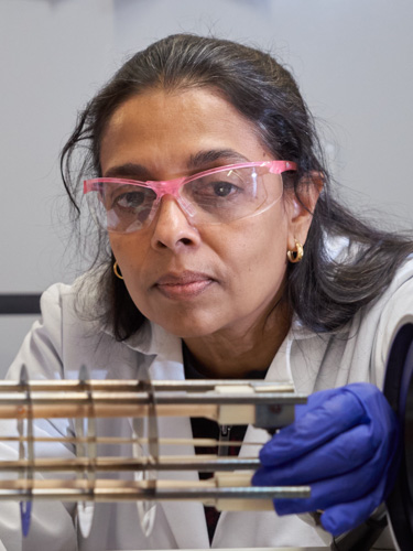 Scientist wearing pink safety glasses and blue gloves examines a metal apparatus in a laboratory.