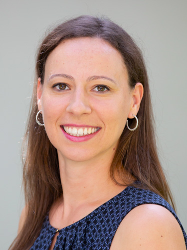 Smiling woman with long brown hair wearing hoop earrings and a sleeveless patterned top against a light gray background.
