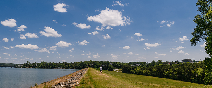 A man walking on a trail on Clemson University's campus with a lake to the left and the football stadium in the background on the right.