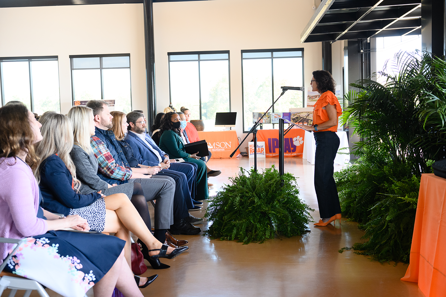 A woman in an orange shirt, navy pants and orange high heels stands at a podium in front of a seated audience.