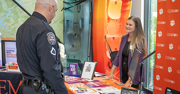 A woman wearing a purple blazer and a man wearing a police uniform talk over a table.