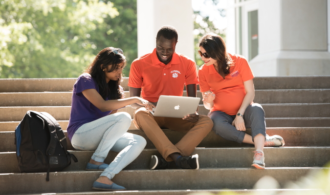 Three employees looking at a laptop together while sitting on steps outside on a sunny day.