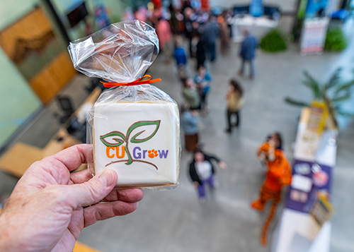 A hand holds a white sugar cookie with the CU Grow logo on it with a room of people blurry in the background.