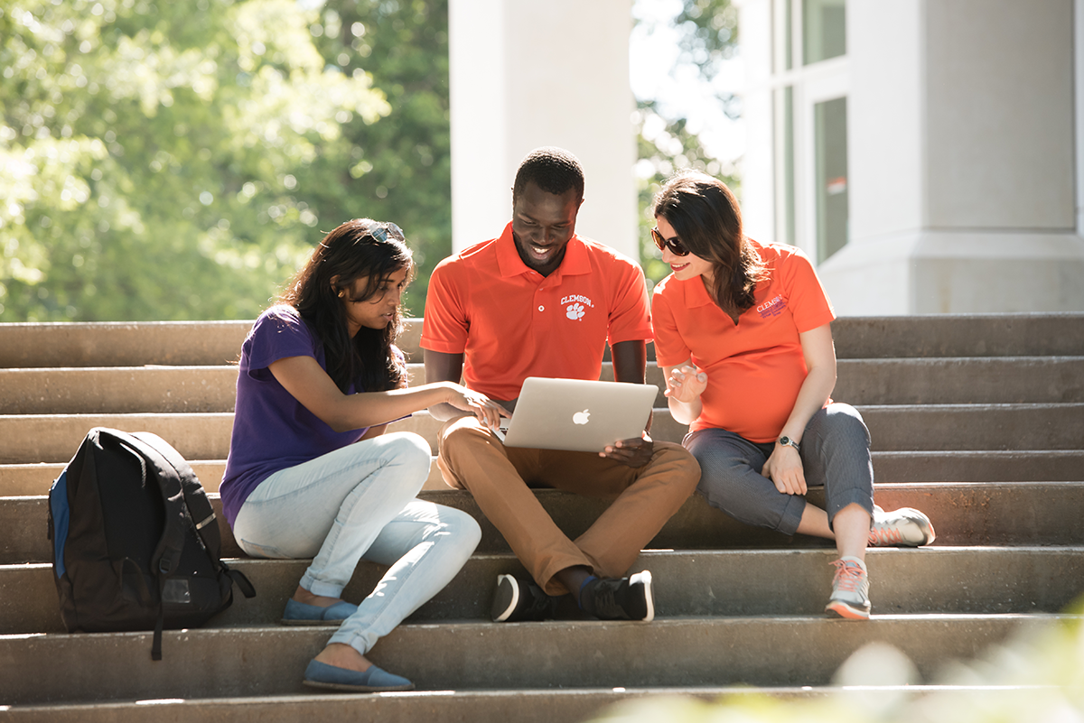 Three employees looking at a laptop together while sitting on steps outside on a sunny day.