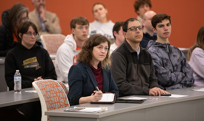 A woman listens to a lecturer and takes notes.