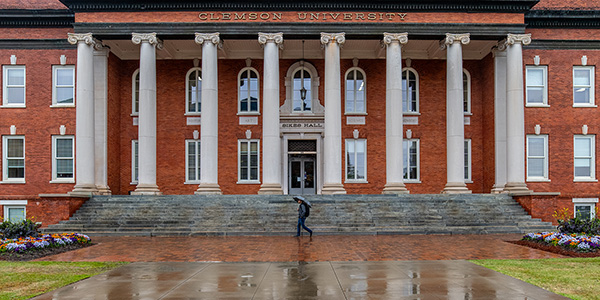 A person walks in front of Sikes Hall holding an umbrella over their head.