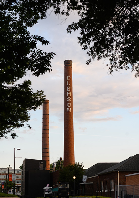 Two brick smoke stacks, one with white bricks spelling "Clemson" down the side stand in the distance behind some trees at the top of the frame.