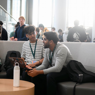 Two students sit on a lounge sofa, talking while one holds a laptop, with a bright, busy atrium in the background.