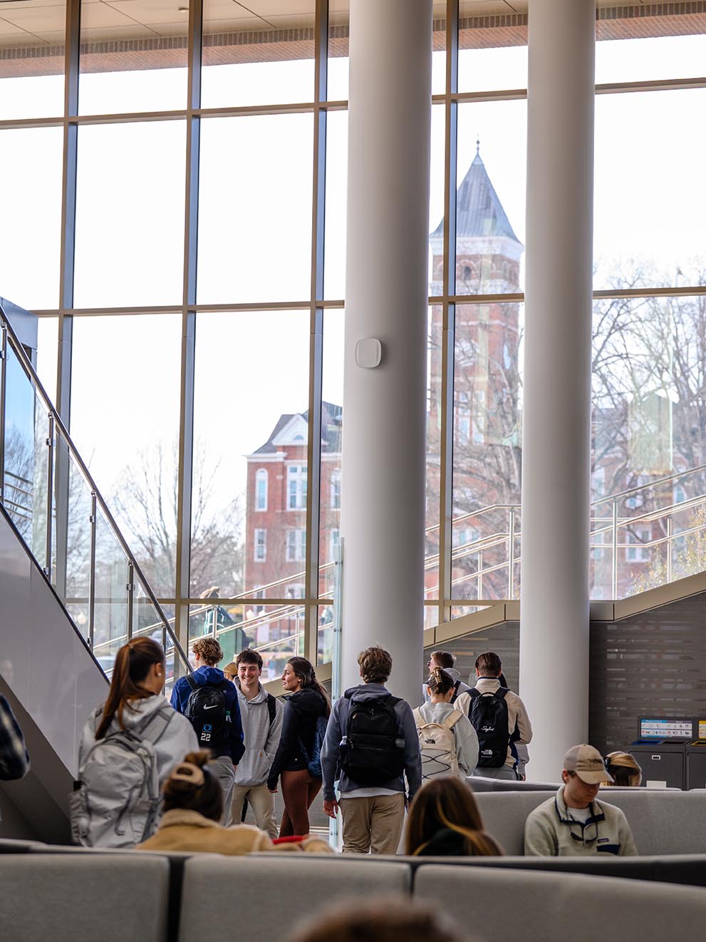Students with backpacks gather and walk through a bright university lobby with tall windows, with a campus building visible outside.