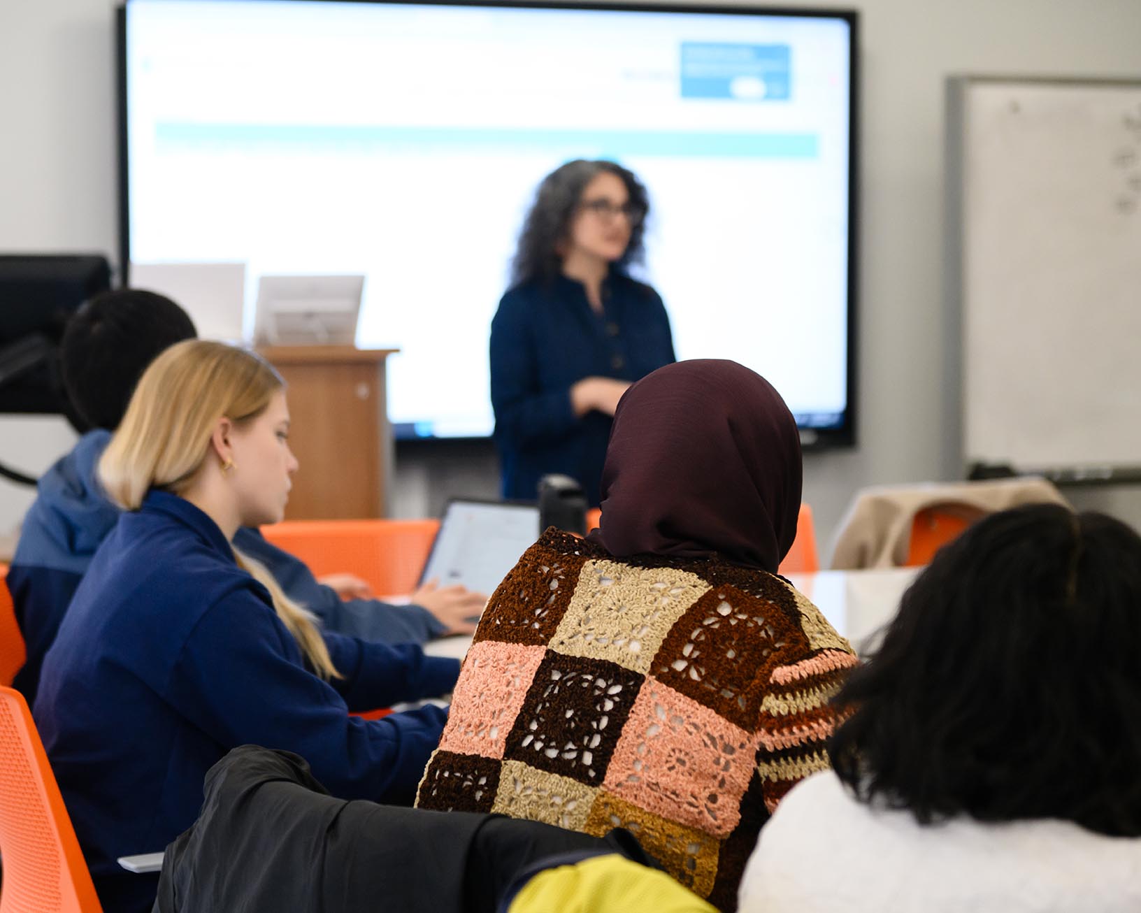 Students sit at desks in a classroom while an instructor presents in front of a large screen.