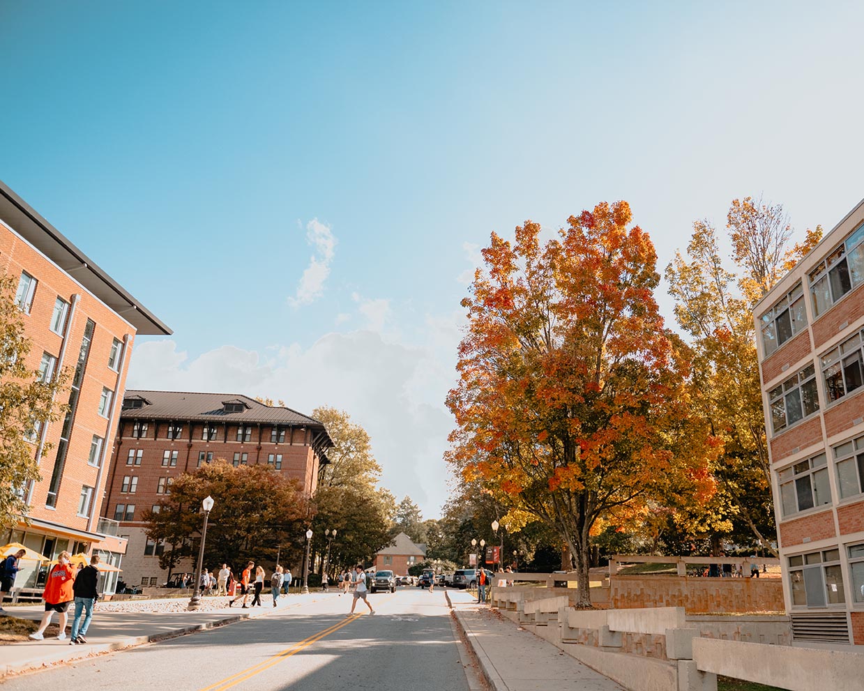 Students walking between brick buildings and trees with bright autumn foliage under a clear blue sky.